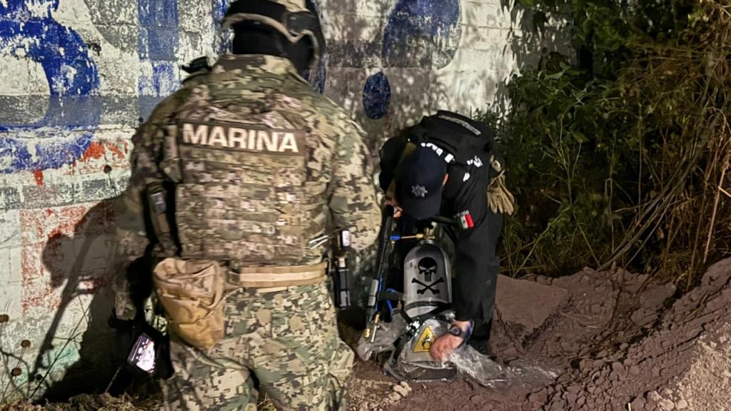 Grupo de oficiales de la Marina y la policía, inspeccionando un área subterránea, con equipo especializado y una mochila que contiene un símbolo de advertencia.