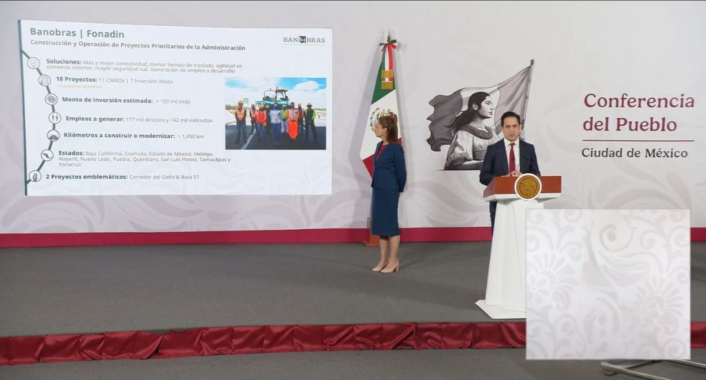 A conference room setup with a podium featuring a microphone and a seal. Two presenters are visible, one speaking while the other stands beside. A national flag is displayed in the background along with a large screen showcasing project details related to infrastructure construction and operation in Mexico.