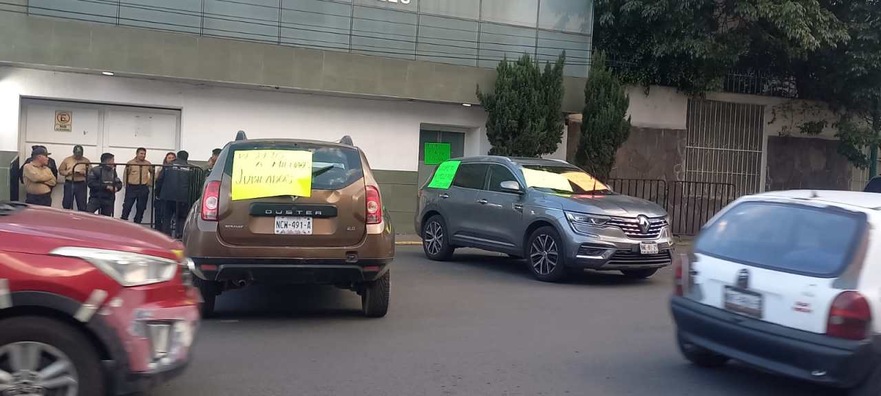 Vehículos estacionados en la calle con carteles de protesta, un grupo de personas observa desde la acera.