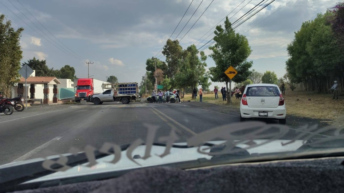Traffic accident scene on a road, featuring a white car and a truck, with several onlookers and debris scattered on the ground.