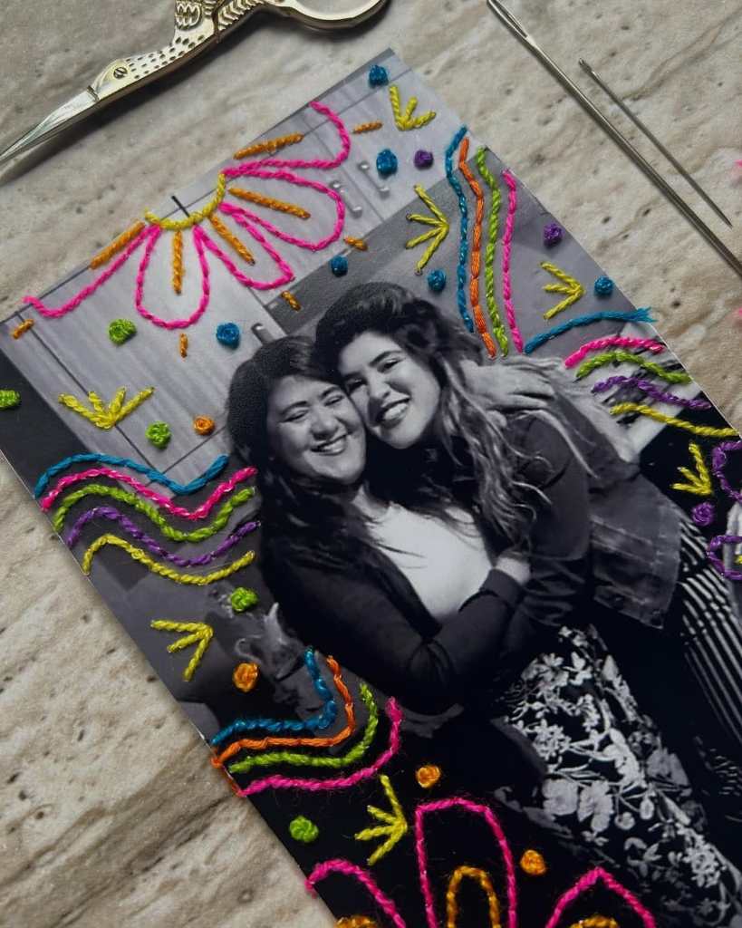 Foto en blanco y negro de dos mujeres sonriendo y abrazándose, decorada con bordados coloridos alrededor.