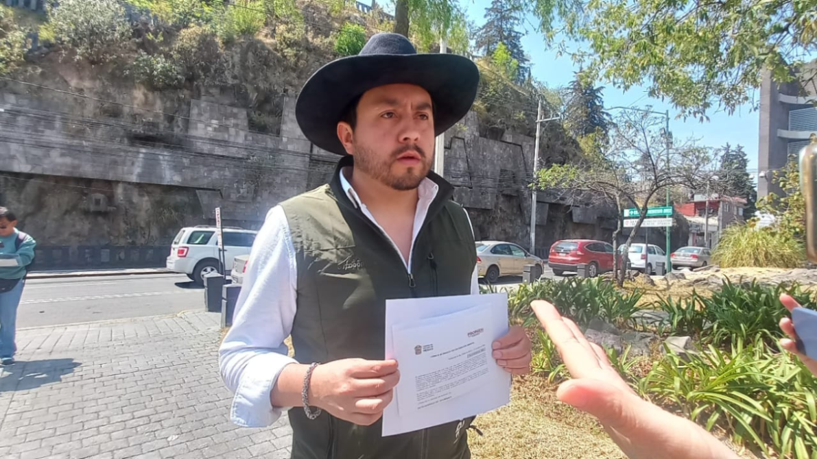 Man wearing a cowboy hat and a green vest stands in an outdoor setting, holding documents, with an expression of concern. In the background, cars and vegetation are visible.