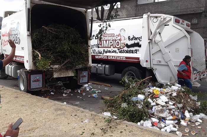 A garbage truck with its rear doors open, revealing a load of branches and trash spilling onto the street, while a person stands nearby, holding a phone.