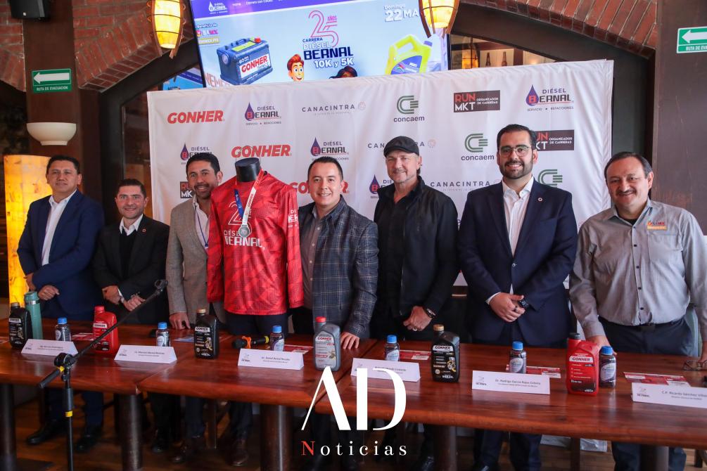 A group of six men dressed in formal attire standing in front of a table displaying branded products and a promotional banner for the Bernal 10K and 5K race.