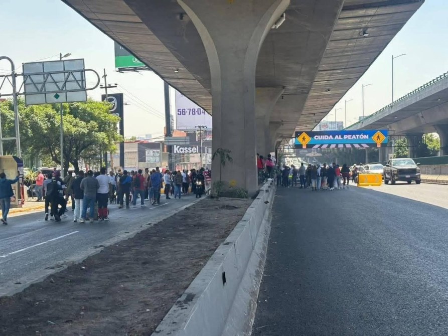 Multitud de personas caminando a un lado de la carretera, con señalización que advierte sobre la precaución al peatón y un puente elevado al fondo.