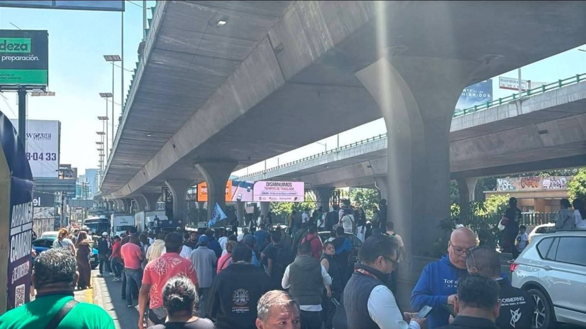 Multitud de personas reunidas bajo un puente, con vehículos estacionados y carteles visibles en el fondo.