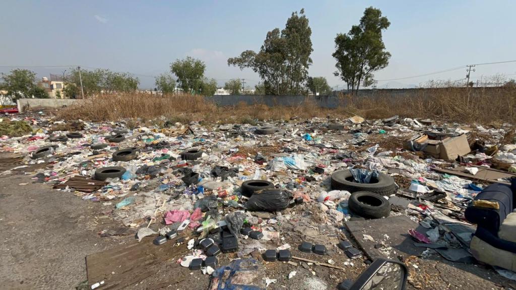 Una gran acumulación de basura y escombros en un terreno baldío, con neumáticos tirados y vegetación seca alrededor.