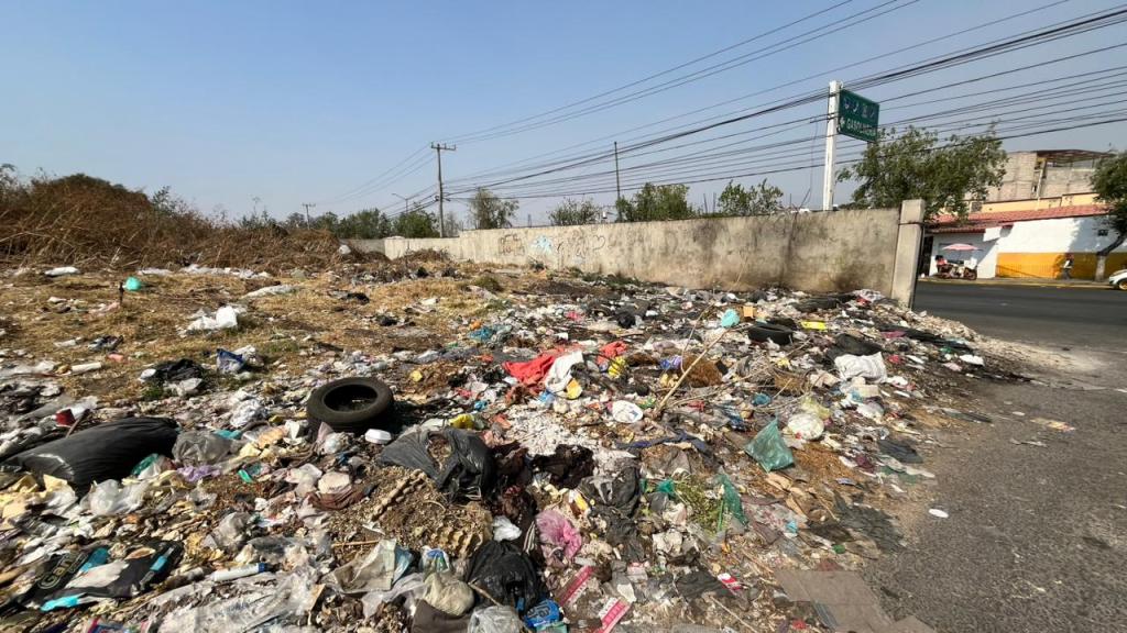 Montón de basura en un terreno baldío junto a una calle, con residuos plásticos y neumáticos.