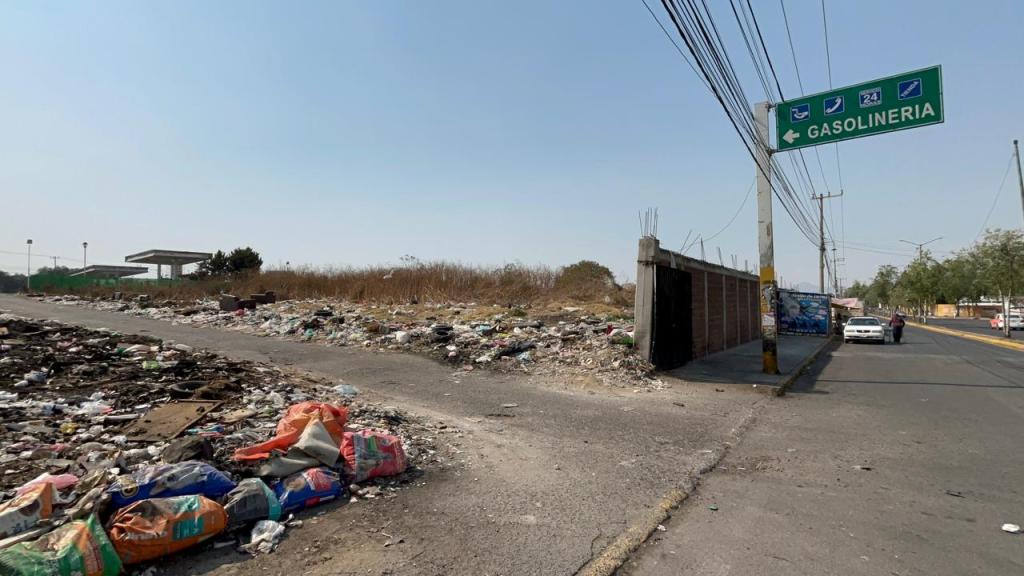 Vista de un camino con basura acumulada a un lado, señal de dirección hacia una gasolinera en la intersección.