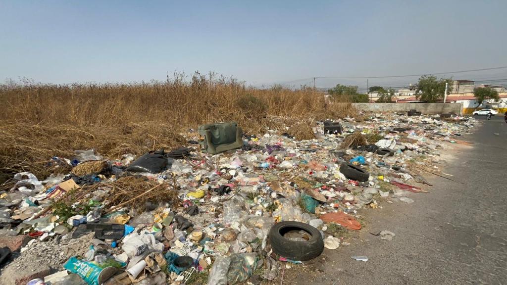 Montón de basura acumulada a un lado de la carretera, con botellas, plásticos y desechos diversos en un área descuidada.