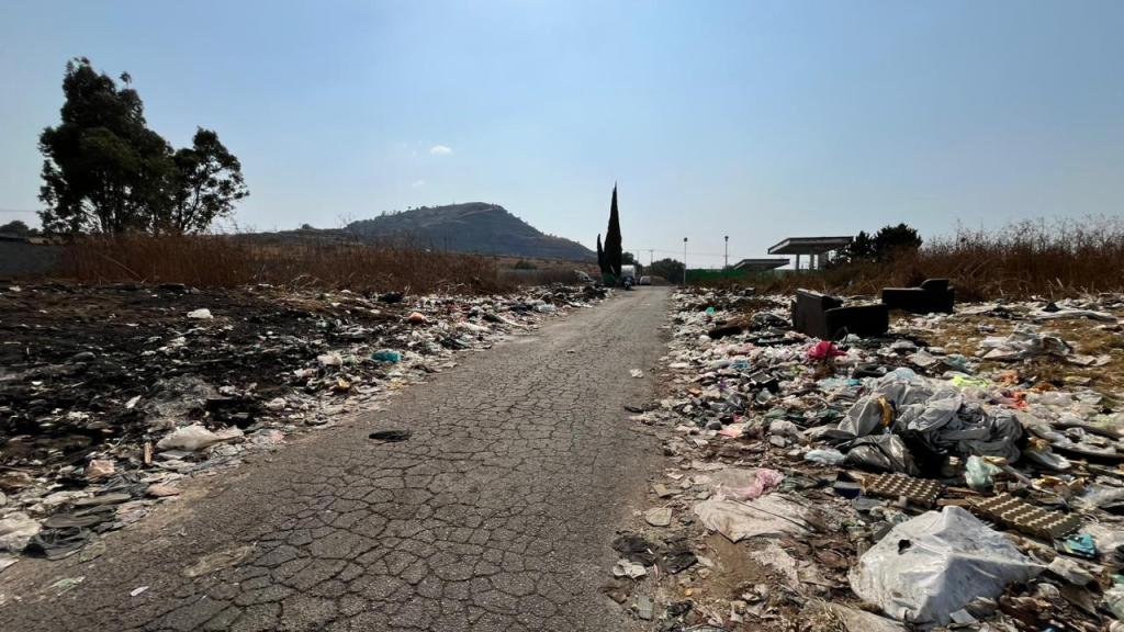 Camino solitario flanqueado por montones de basura, con un paisaje montañoso al fondo y arbustos secos a los lados.
