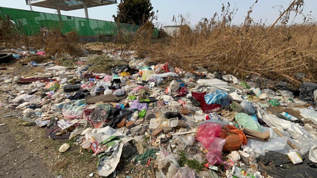Montón de basura y desechos en un área desierta, con vegetación seca al fondo.