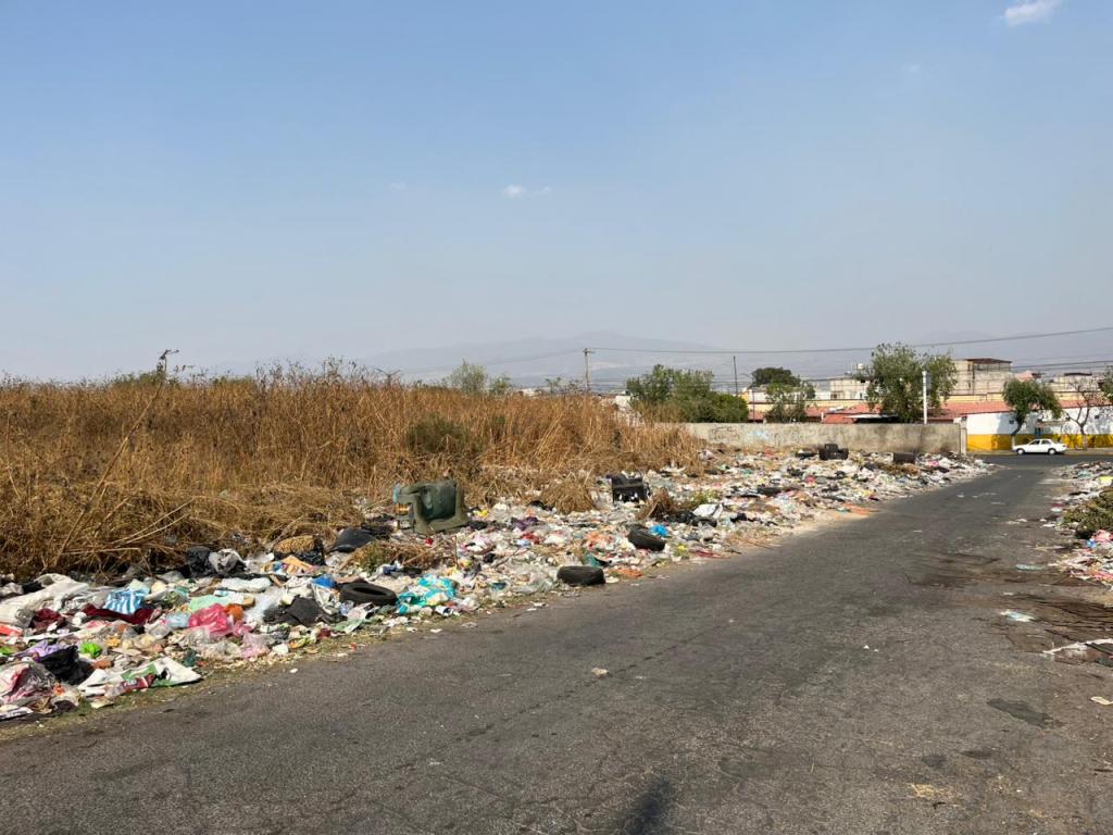 Vista de un área urbana contaminada con basura y escombros, con vegetación seca y un camino pavimentado al fondo.