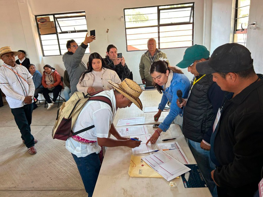 Un grupo de personas en una sala durante un proceso electoral, con un hombre de sombrero firmando documentos mientras otros observan.