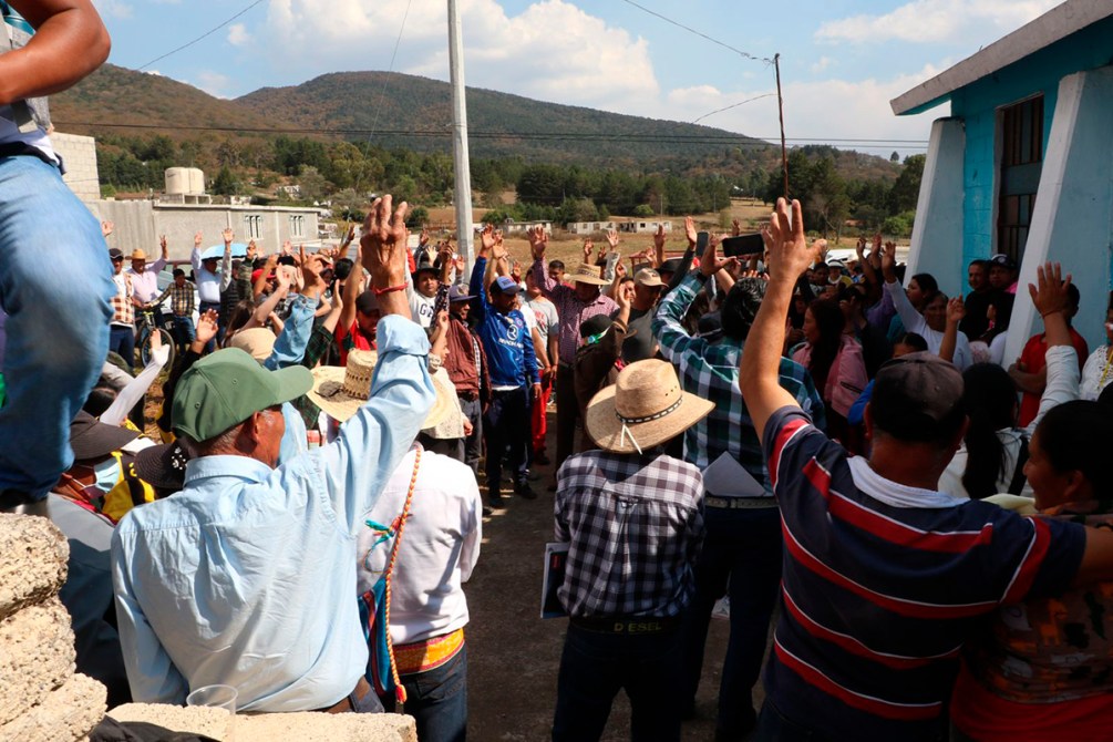 Multitud de personas reunidas al aire libre, con muchas manos levantadas, en un ambiente comunitario en una zona rural.