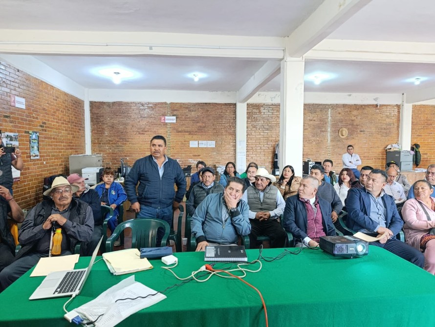Reunión en un salón con un grupo de personas, algunas de ellas sentadas alrededor de una mesa verde. Un hombre está de pie hablando mientras los demás lo observan.