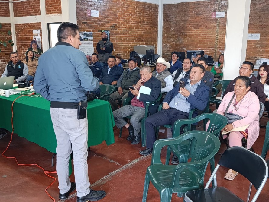 Reunión de un grupo de personas sentadas en sillas, escuchando a un orador. Se observa un escritorio con una mesa verde y equipos electrónicos en el fondo.