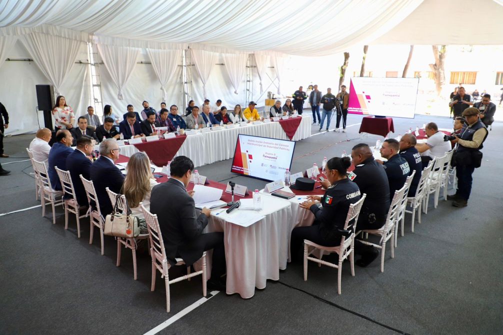 A large group of officials and law enforcement personnel participating in a meeting under a tent, with a presentation screen in the foreground displaying information.