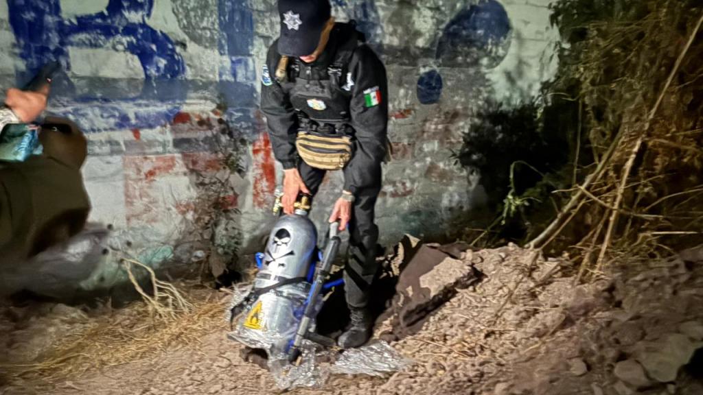 Un oficial de policía manipula un dispositivo en un área sombría, con graffiti de fondo.