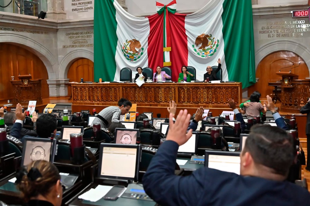 Sesion en un recinto legislativo con diputados levantando la mano en votación, en un ambiente formal.