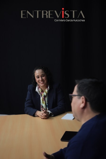 Una mujer sonriente vestida de traje conversa frente a un hombre en una mesa, con un fondo oscuro, durante una entrevista.