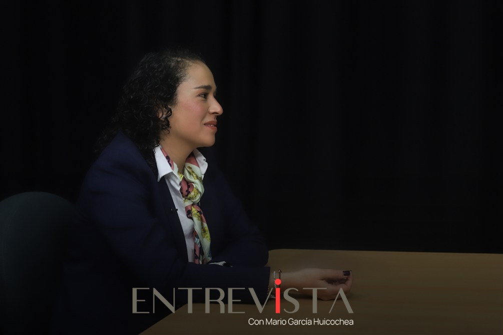 Una mujer sentada en una mesa, mirando hacia un lado, con una expresión amable. Lleva un blazer azul y una bufanda colorida. Fondo oscuro.