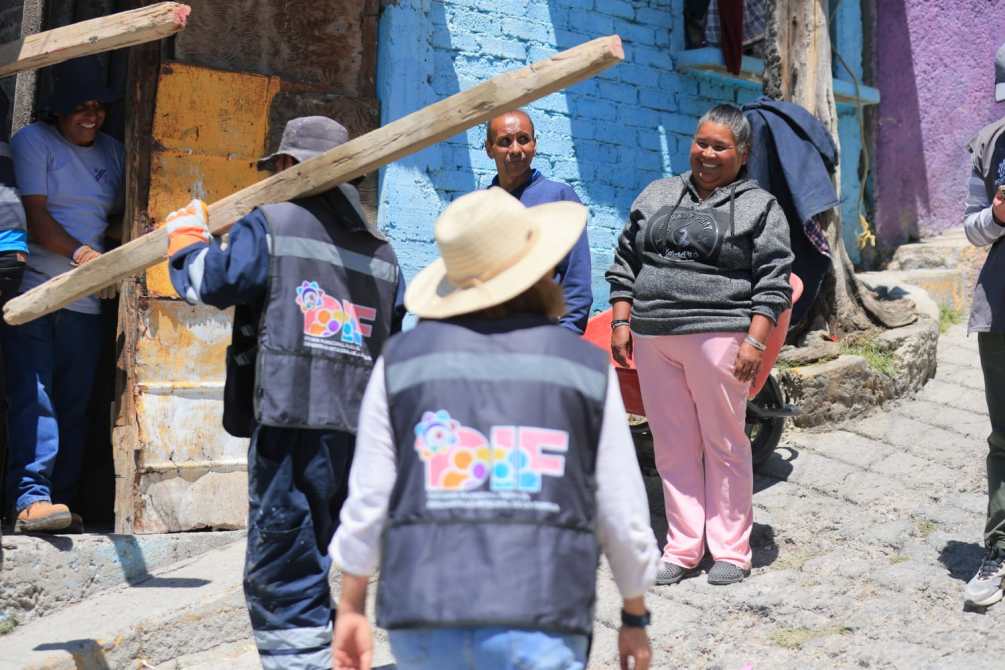 A group of people interacting in a community setting, with one person carrying a wooden beam while others chat and smile. Colorful buildings are visible in the background.