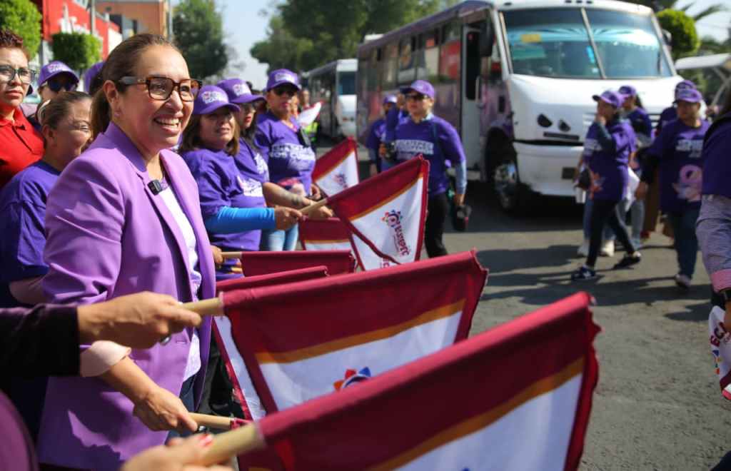 Una mujer sonriente con gafas, vestida de morado, sostiene una bandera mientras se encuentra al frente de un grupo de mujeres que también sostienen banderas. Hay un autobús y árboles de fondo.