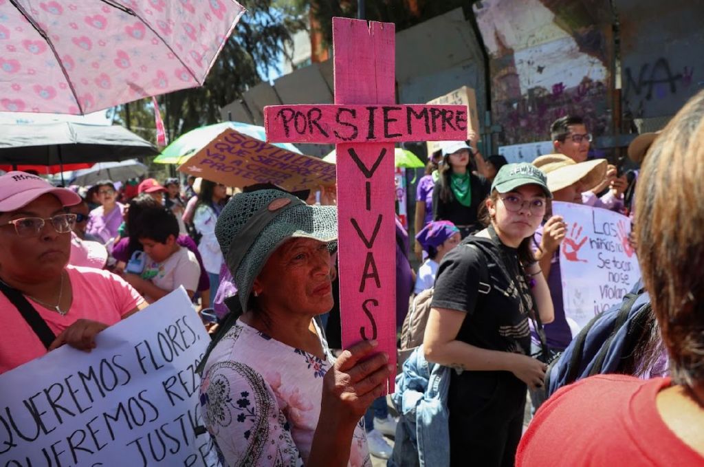 Manifestación con mujeres sosteniendo pancartas, incluyendo una cruz rosa que dice 'POR SIEMPRE VIVAS'.