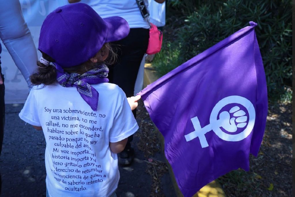 Niña sosteniendo una bandera morada con el símbolo feminista, vistiendo una camiseta con un mensaje sobre ser sobreviviente y no víctima.