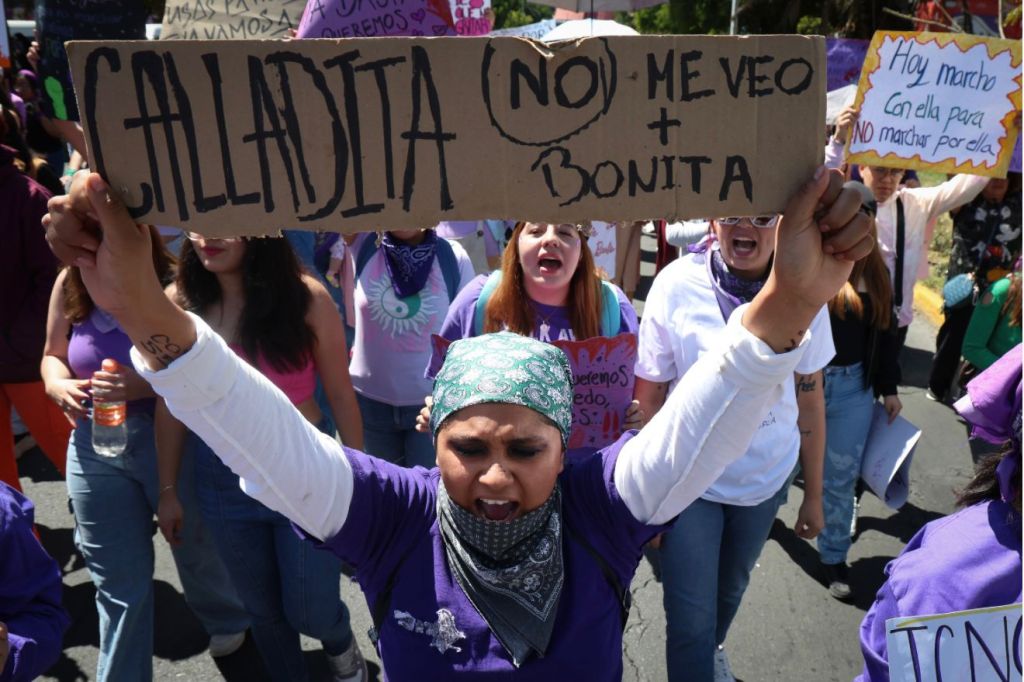 Una mujer sostiene un cartel que dice 'CALLADITA NO ME VEO BONITA' mientras participa en una marcha con un grupo de personas, todos vestidos con ropa morada y sosteniendo pancartas.