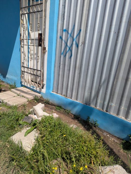 Puerta de entrada con rejas en una pared azul, junto a un cercado de metal con pintura azul y una marca azul en forma de cruz, rodeada de hierba y piedras en el suelo.