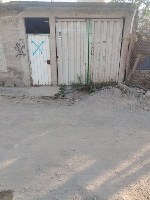 Entrada de un cobertizo con puertas de metal, una de ellas marcada con una 'X'. El entorno es un camino de tierra con algunas plantas.