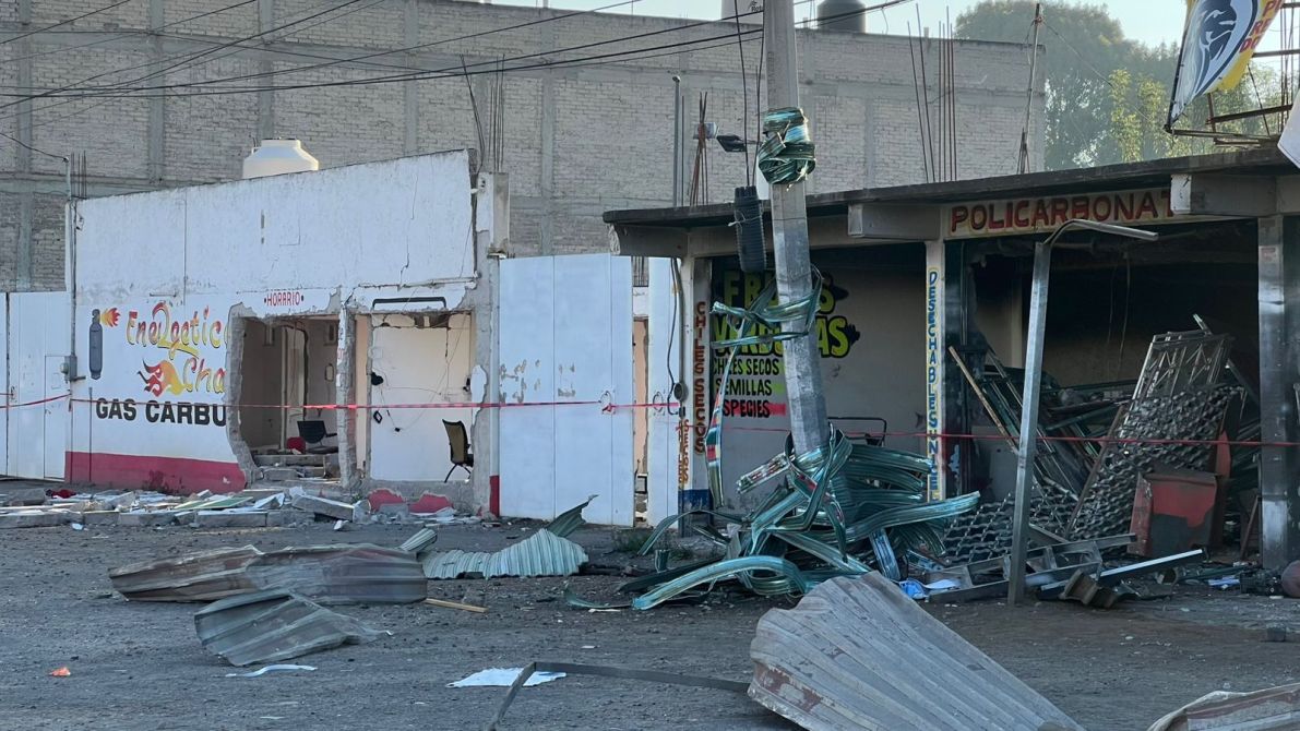Vista de un área comercial dañada, con paredes agrietadas y escombros en el suelo. Se observan señales de advertencia y restos de estructuras de metal en la escena.