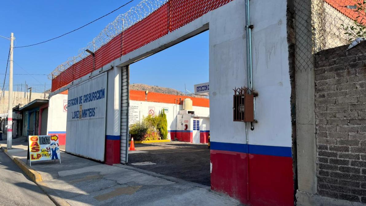 Entrada de una instalación con paredes blancas y rojas, cercada con alambre de púas, y un cartel de gasolinería en la acera.