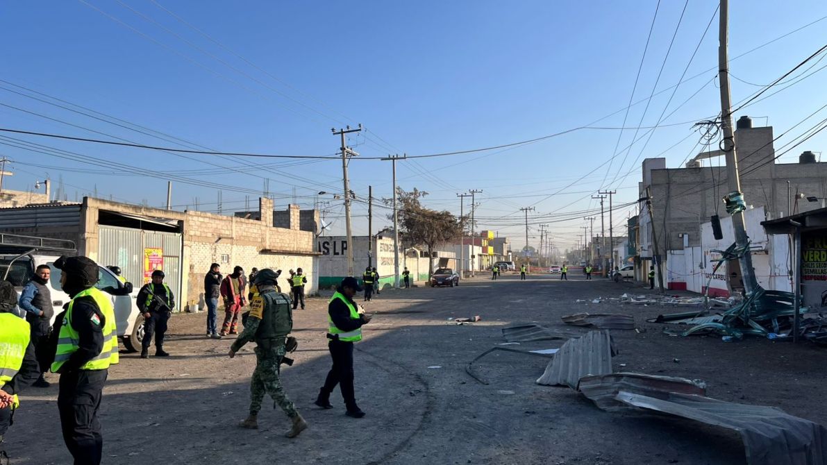 Escena de una calle con presencia de policías y militares, con edificios de ladrillo y cables eléctricos visibles, y escombros en el suelo.
