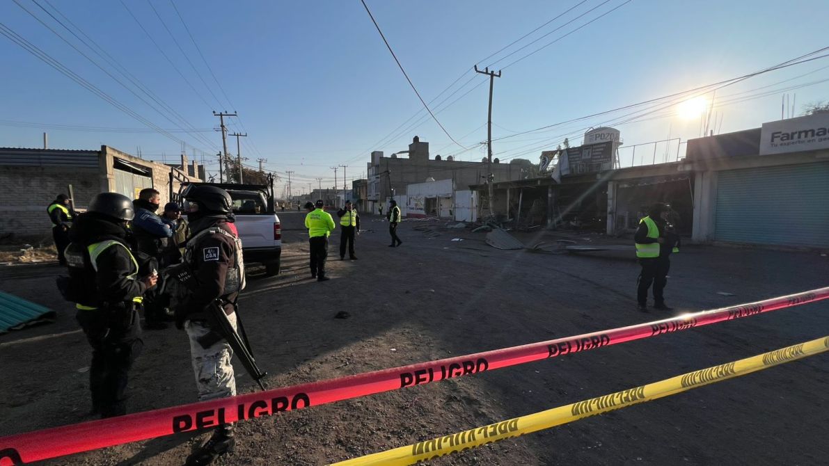 Escena de intervención policial en una calle con presencia de agentes y vehículos, cercada con cintas de advertencia y edificios dañados al fondo.