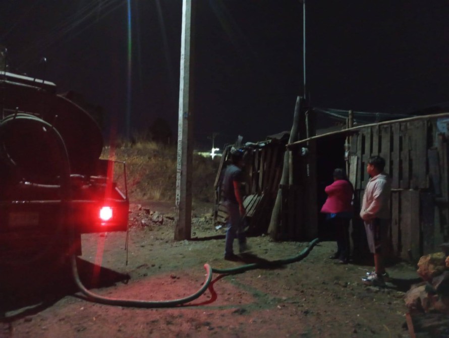 Three individuals standing near a makeshift wooden structure at night, with a truck emitting red light and a hose running towards the building.