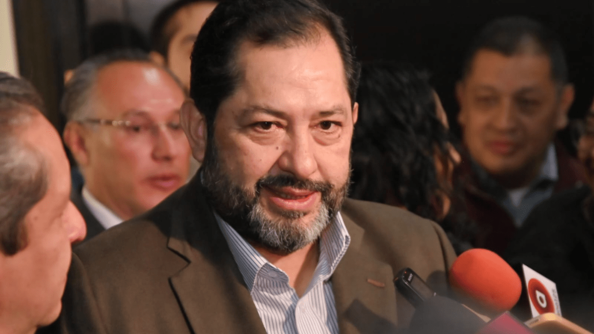 Hombre de mediana edad con barba y chaqueta, hablando a los medios en una conferencia, rodeado de otras personas y cámaras.