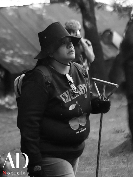 Una mujer con sombrero y sudadera caminando en un entorno natural, sosteniendo un bastón.