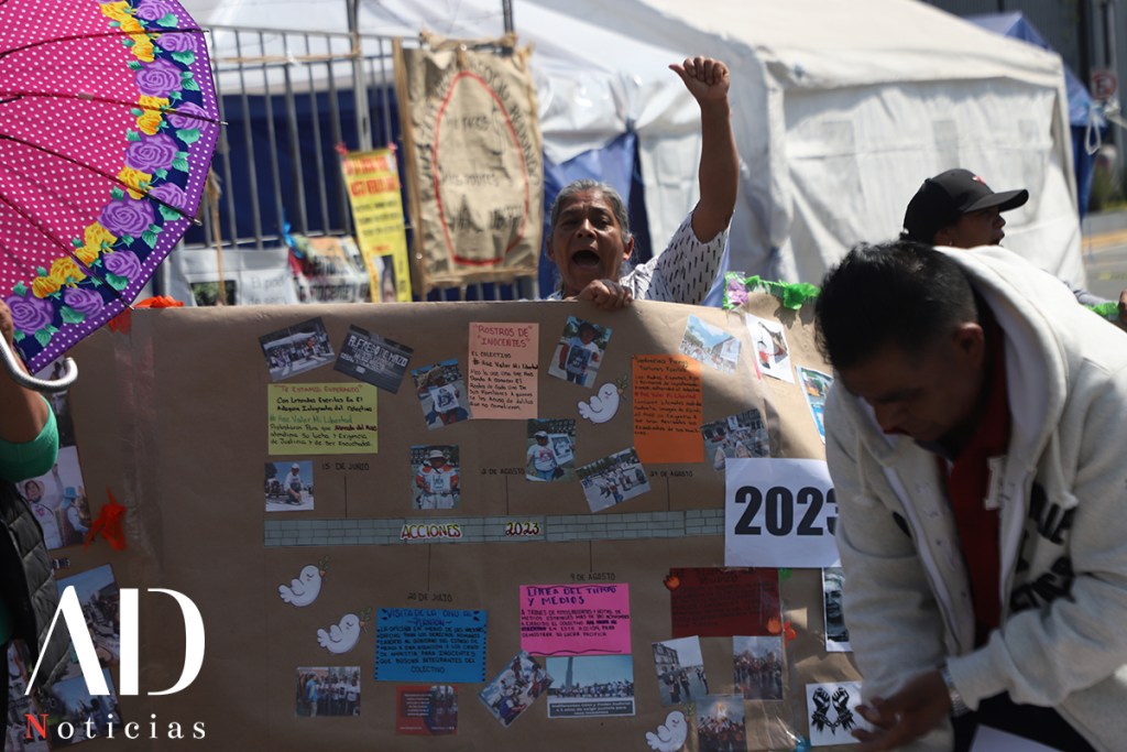 A woman holding a colorful umbrella and shouting in a demonstration, with a poster displaying text and photos behind her, while another man in a sweater is shown nearby.