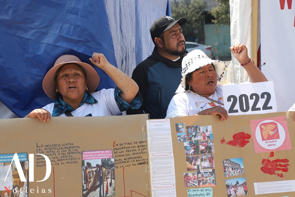 Three individuals participating in a protest, with one woman holding a sign that reads '2022'. They are expressing their message passionately in front of a decorated cardboard display about social issues.