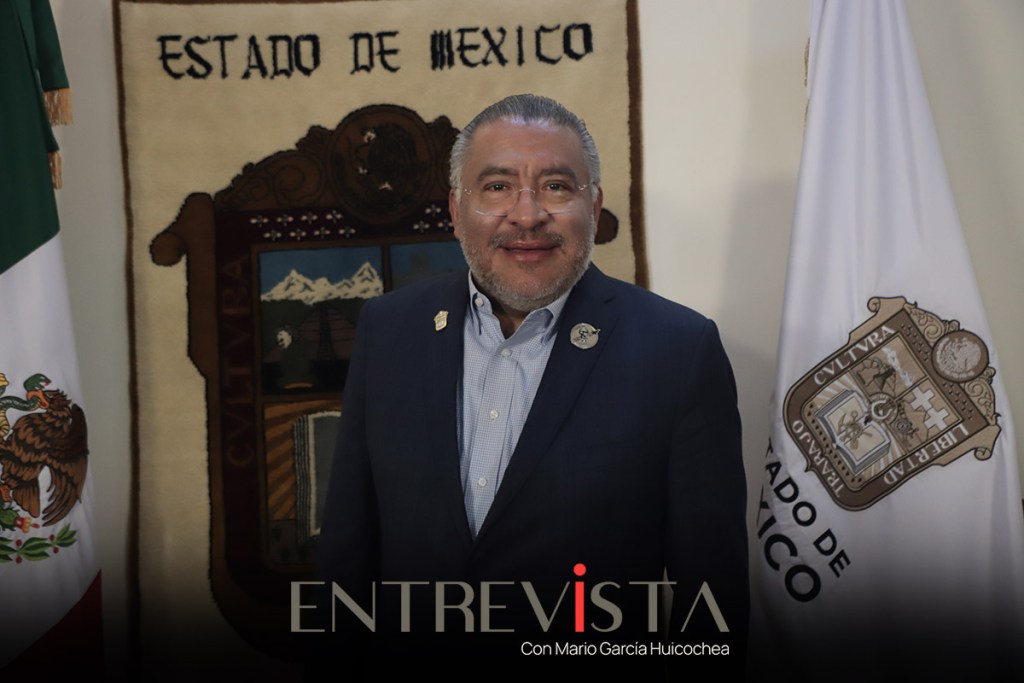 Hombre sonriendo frente a un fondo con banderas y un emblema del Estado de México.