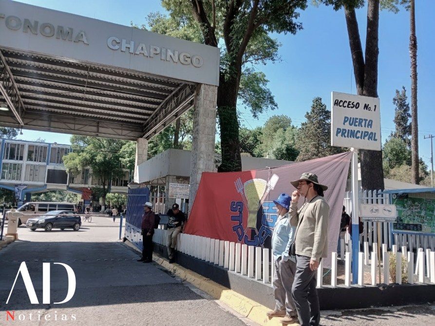 Entrada principal de la Universidad Autónoma Chapingo, con dos personas de pie frente a una bandera y un letrero que indica acceso número 1.