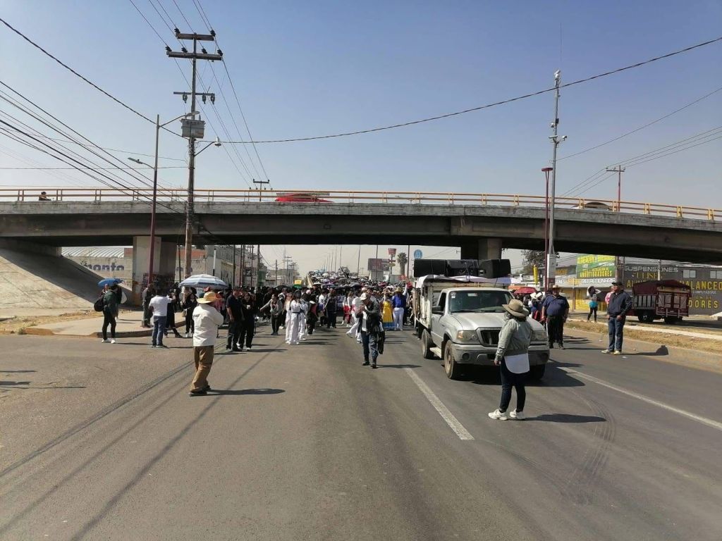 A large crowd of people marching down the street under an overpass, with various vehicles present and commercial buildings visible in the background.