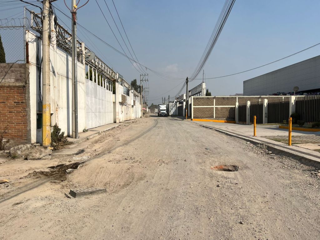 Vista de una calle de tierra en un área industrial, con edificios y muros a ambos lados. Se observan postes de luz y cables eléctricos.