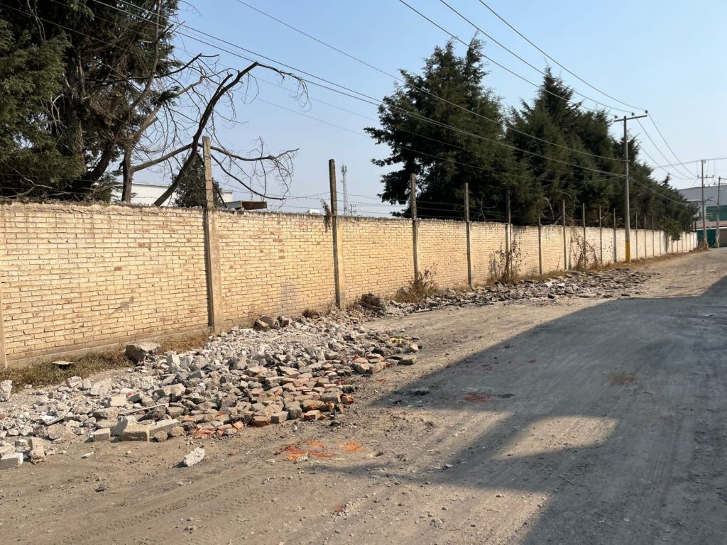 Calle de tierra con escombros a un lado, cercada por un muro de ladrillos y árboles al fondo.