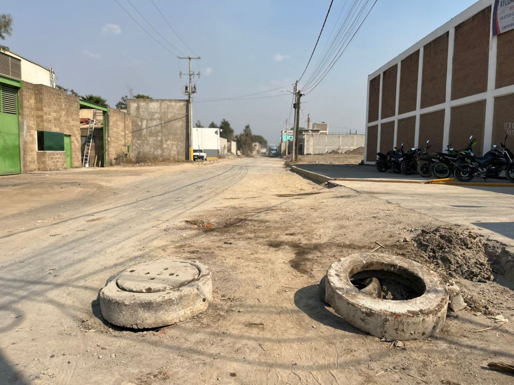 Vista de una calle desierta con pavimento agrietado y dos registros de alcantarilla en el primer plano, rodeada de edificios y un estacionamiento de motocicletas.