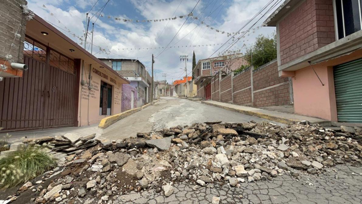 A damaged street with a pile of rubble blocking the roadway, surrounded by residential buildings under a cloudy sky.