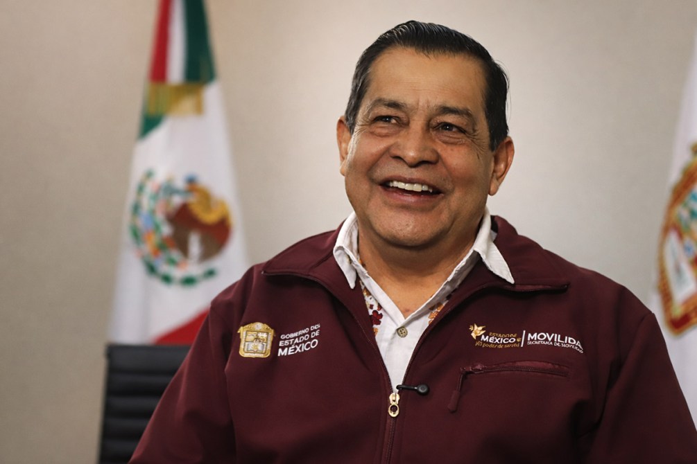 Hombre sonriente en una camisa formal y chaqueta con el logotipo del Gobierno del Estado de México, sentado frente a un fondo con banderas de México.